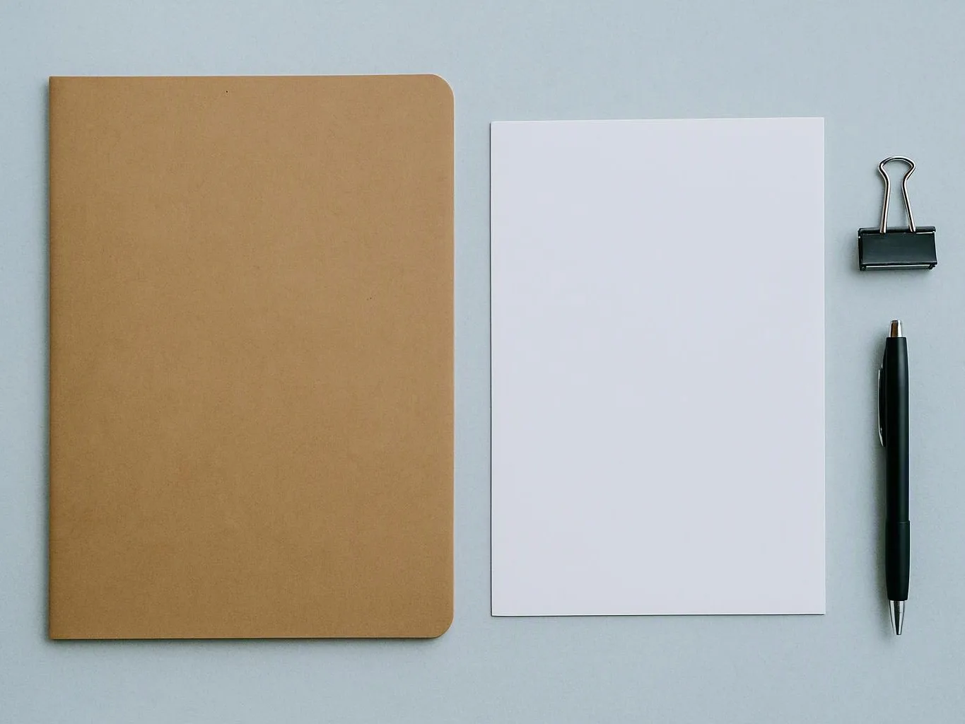 A brown notebook, blank white paper, binder clip, and black pen rest on a pale gray desk
