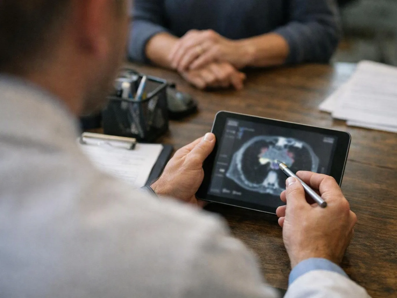 A person studies a medical scan on a tablet with a stylus at a desk across from another person