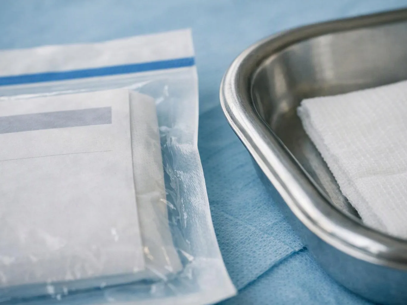 A sealed plastic pouch and folded paper sit beside a metal tray holding white gauze on a blue surgical drape