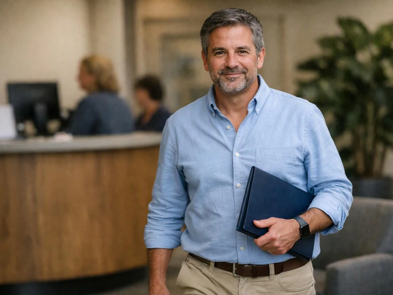 A smiling man in a light blue shirt holds a dark folder in an office lobby