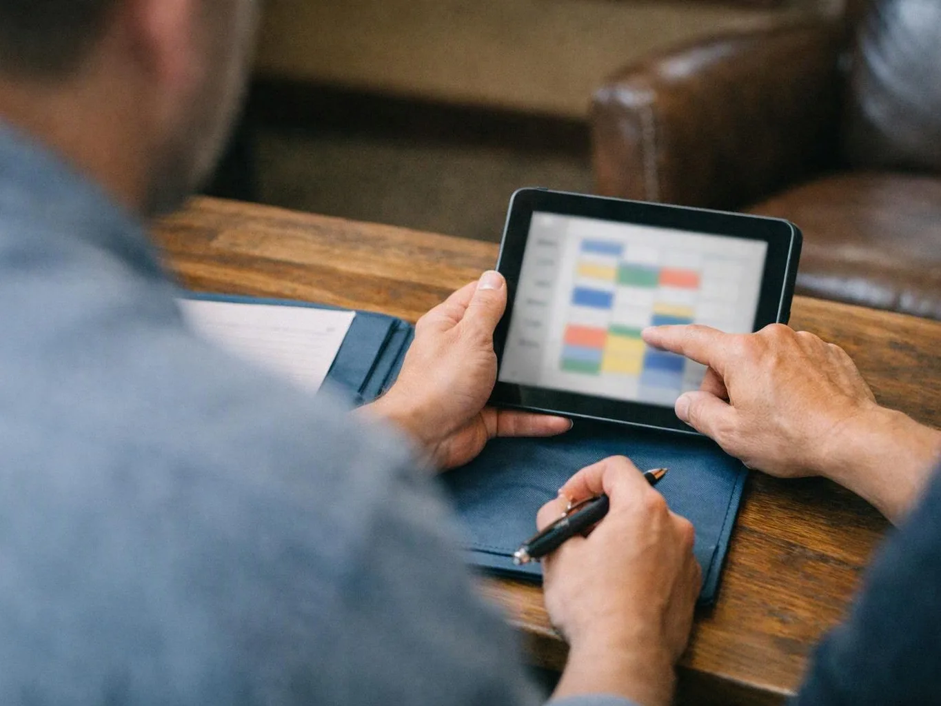 Two people study a tablet with a colorful calendar on a wooden table indoors